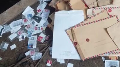 Foto close-up tumpukan amplop coklat lamaran kerja berserakan di pinggir jalan aspal, beberapa terbuka memperlihatkan dokumen di dalamnya.