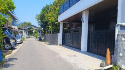 Rumah putih dengan pagar besi dan tanaman hias di depan, tampak sepi.